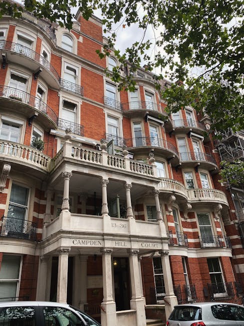 A narrow, cobbled residential street with brick and stucco buildings on the right side, featuring multiple large sash windows and a black drainpipe running down the facade. Potted plants and greenery