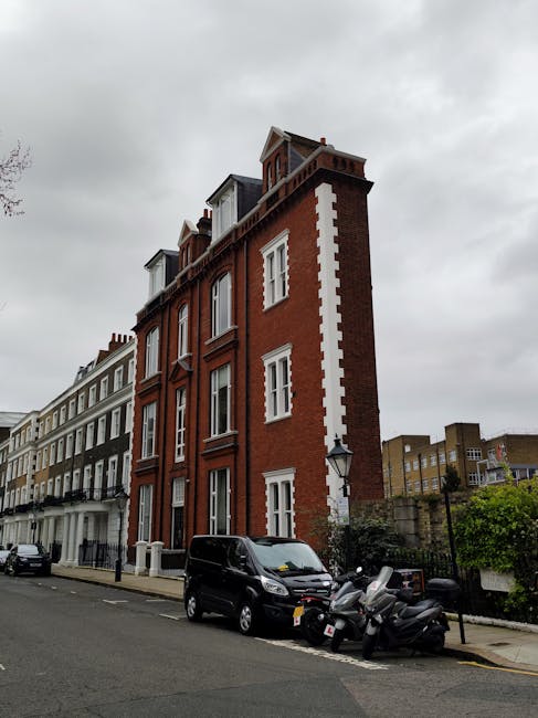 Exterior view of a red-brick Victorian-style building located on a residential street in West Kensington, London, with tall windows, decorative iron railings on small balconies, and a white awning ove